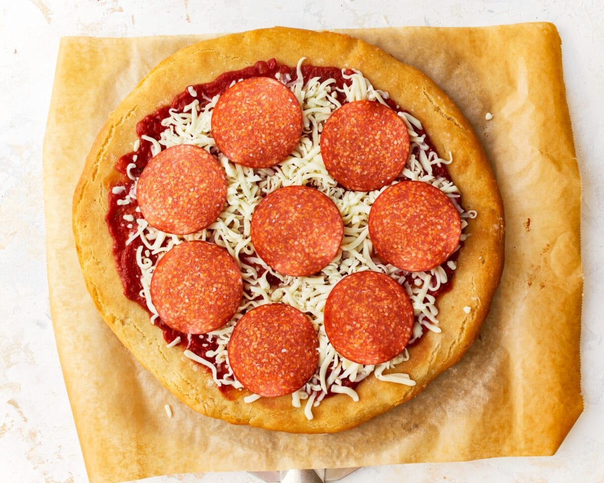 A partially assembled gluten-free pepperoni pizza with tomato sauce, shredded mozzarella, and evenly spaced pepperoni slices on a baked crust, ready to go into the oven.