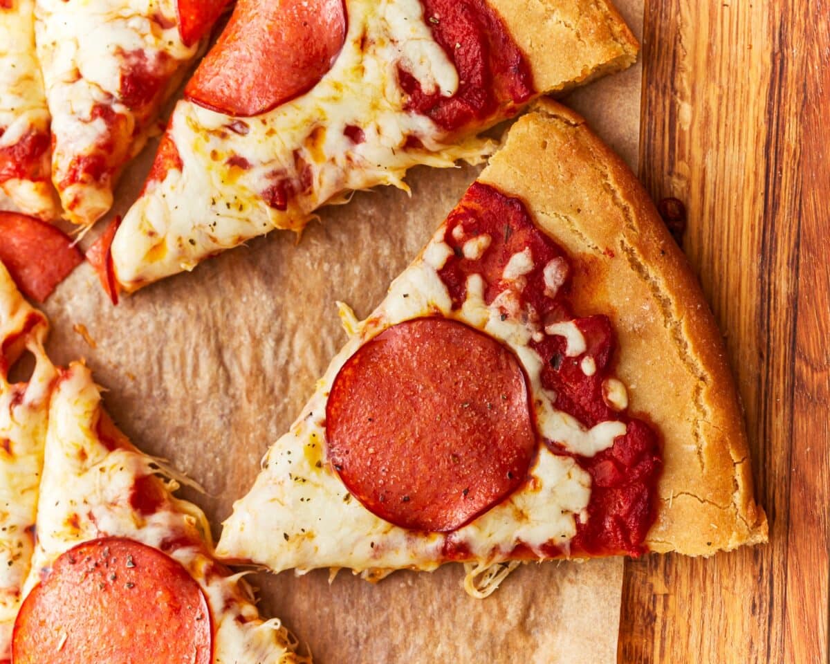 A close-up view of sliced gluten-free pepperoni pizza on a wooden board, with one slice pulled away showing the thick crust and melted cheese.