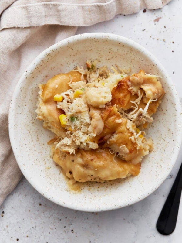 A serving of gluten-free chicken and dumplings casserole on a white speckled plate with visible chunks of biscuit topping, shredded chicken, corn, and creamy sauce.