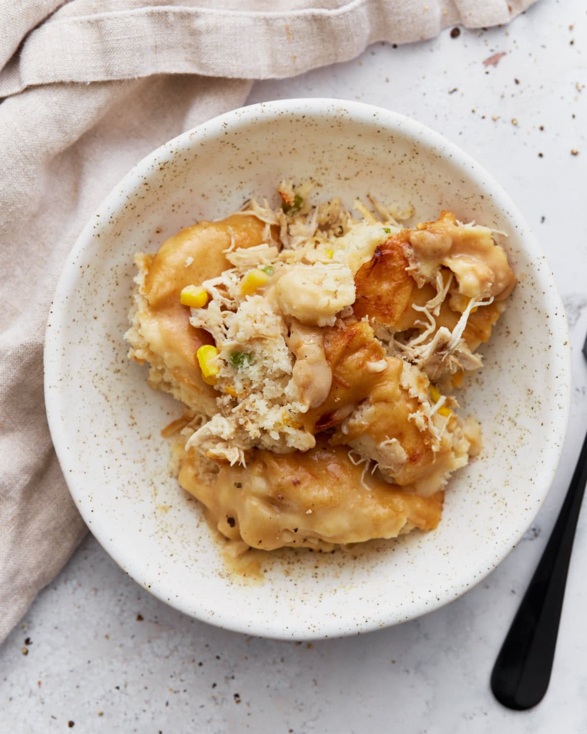 A serving of gluten-free chicken and dumplings casserole on a white speckled plate with visible chunks of biscuit topping, shredded chicken, corn, and creamy sauce.