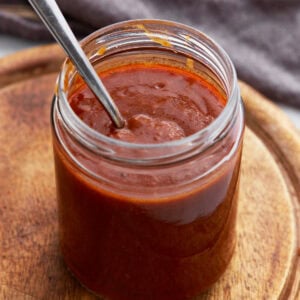 A glass jar filled with rich, dark red enchilada sauce with a spoon inside, sitting on a wooden board.
