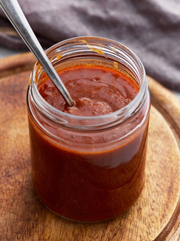 A glass jar filled with rich, dark red enchilada sauce with a spoon inside, sitting on a wooden board.