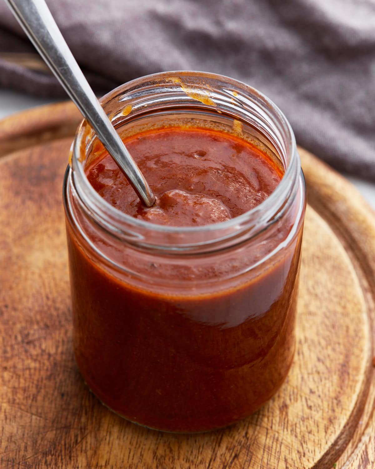 A glass jar filled with rich, dark red enchilada sauce with a spoon inside, sitting on a wooden board.