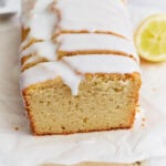 Sliced gluten-free iced lemon loaf on parchment paper with a thick white glaze dripping over the edges.