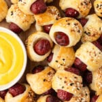 Close-up of golden-brown gluten-free pigs in a blanket topped with everything bagel seasoning, served next to a bowl of yellow mustard.