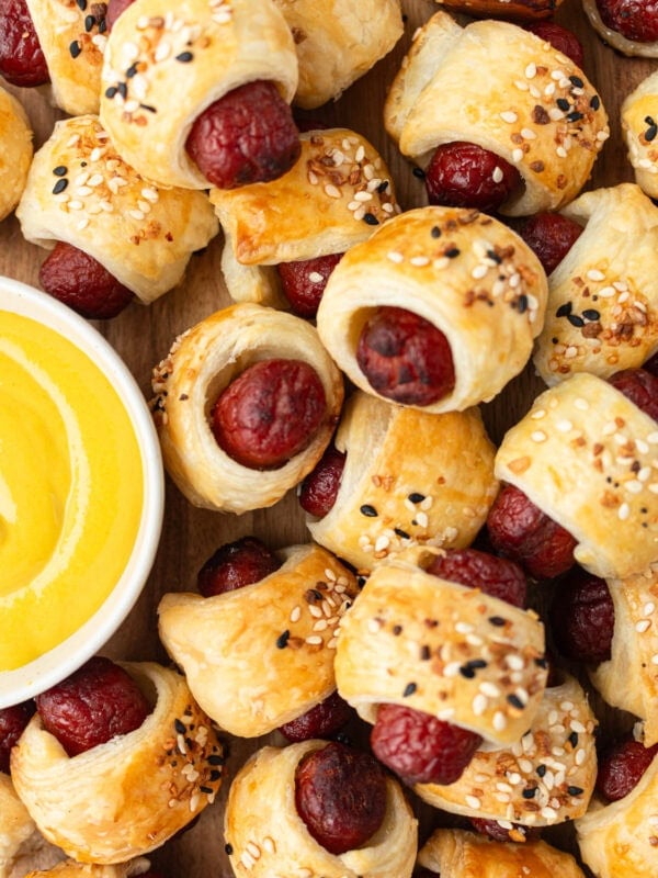 Close-up of golden-brown gluten-free pigs in a blanket topped with everything bagel seasoning, served next to a bowl of yellow mustard.