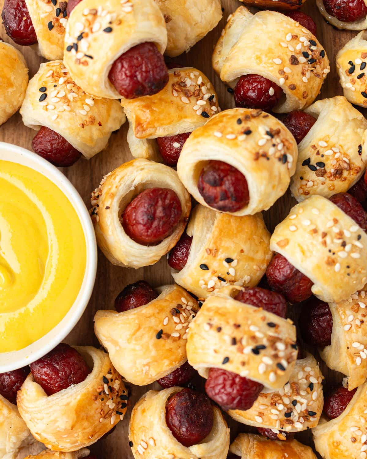 Close-up of golden-brown gluten-free pigs in a blanket topped with everything bagel seasoning, served next to a bowl of yellow mustard.