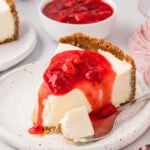 A slice of gluten-free no-bake cheesecake topped with strawberry sauce on a speckled plate, with a bite taken and a spoon beside it.