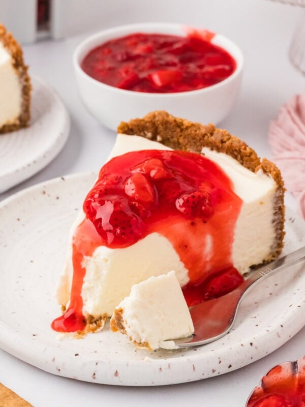 A slice of gluten-free no-bake cheesecake topped with strawberry sauce on a speckled plate, with a bite taken and a spoon beside it.