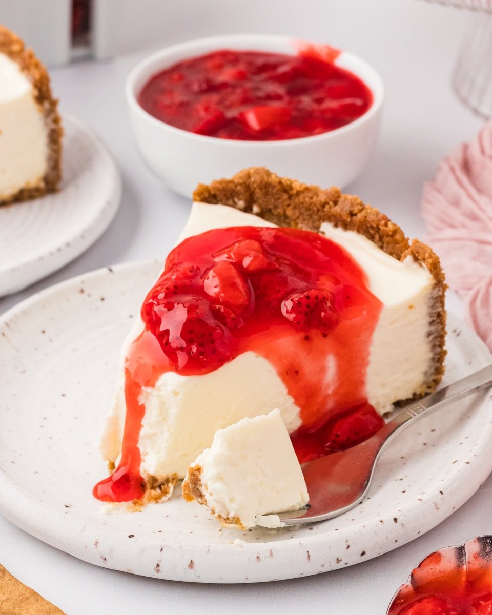 A slice of gluten-free no-bake cheesecake topped with strawberry sauce on a speckled plate, with a bite taken and a spoon beside it.