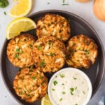 Overhead view of five golden-brown gluten-free crab cakes served on a dark plate with lemon wedges and a bowl of creamy dipping sauce.