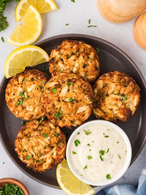 Overhead view of five golden-brown gluten-free crab cakes served on a dark plate with lemon wedges and a bowl of creamy dipping sauce.