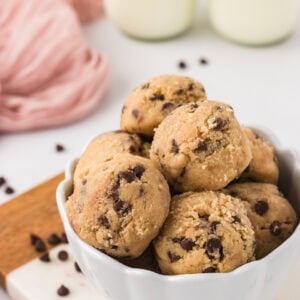 Bowl filled with scoops of gluten-free edible chocolate chip cookie dough, surrounded by scattered mini chocolate chips and glasses of milk in the background.