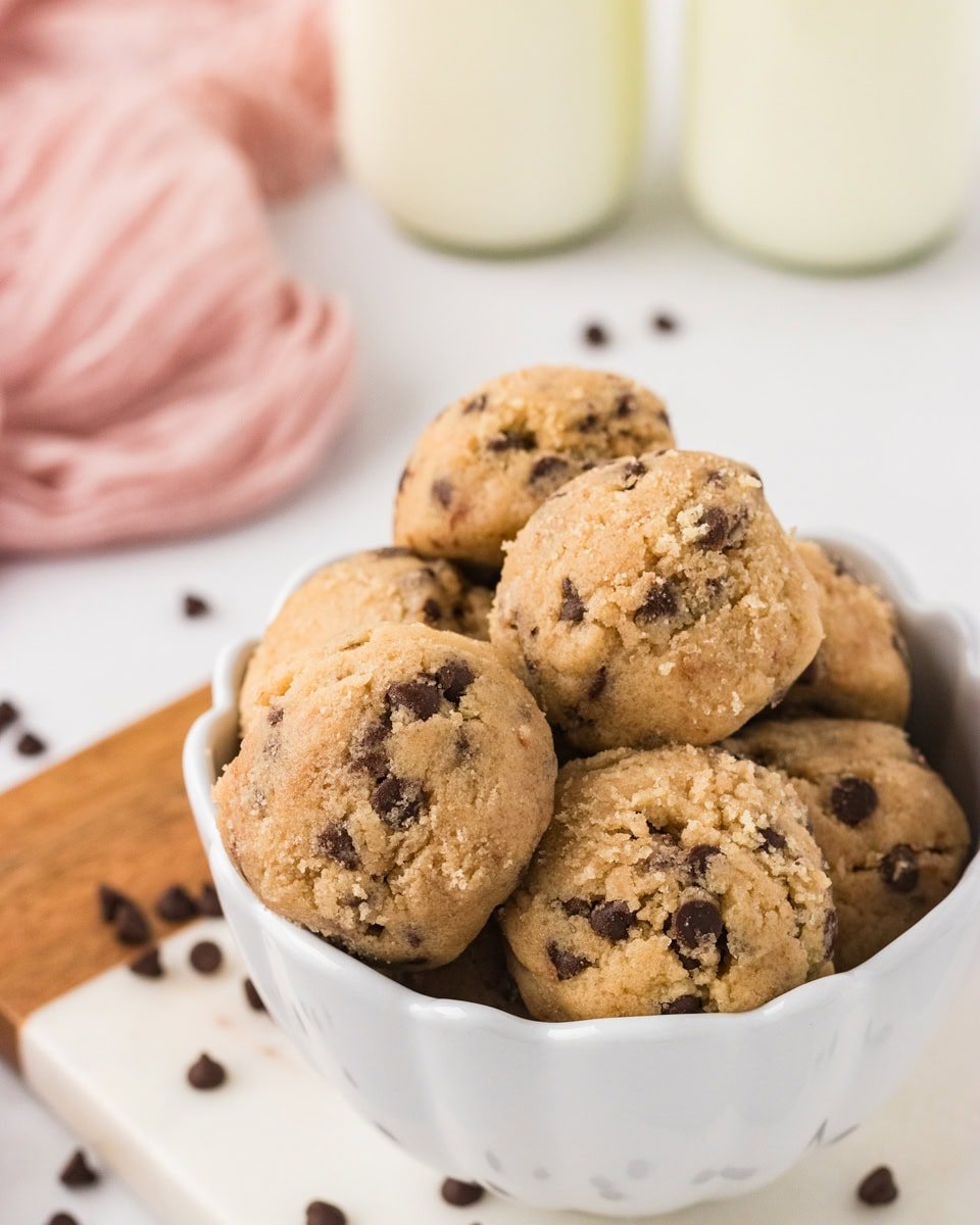 Bowl filled with scoops of gluten-free edible chocolate chip cookie dough, surrounded by scattered mini chocolate chips and glasses of milk in the background.