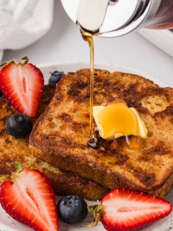 Two slices of golden-brown gluten-free French toast topped with a pat of butter and fresh berries, with maple syrup being poured over the top.