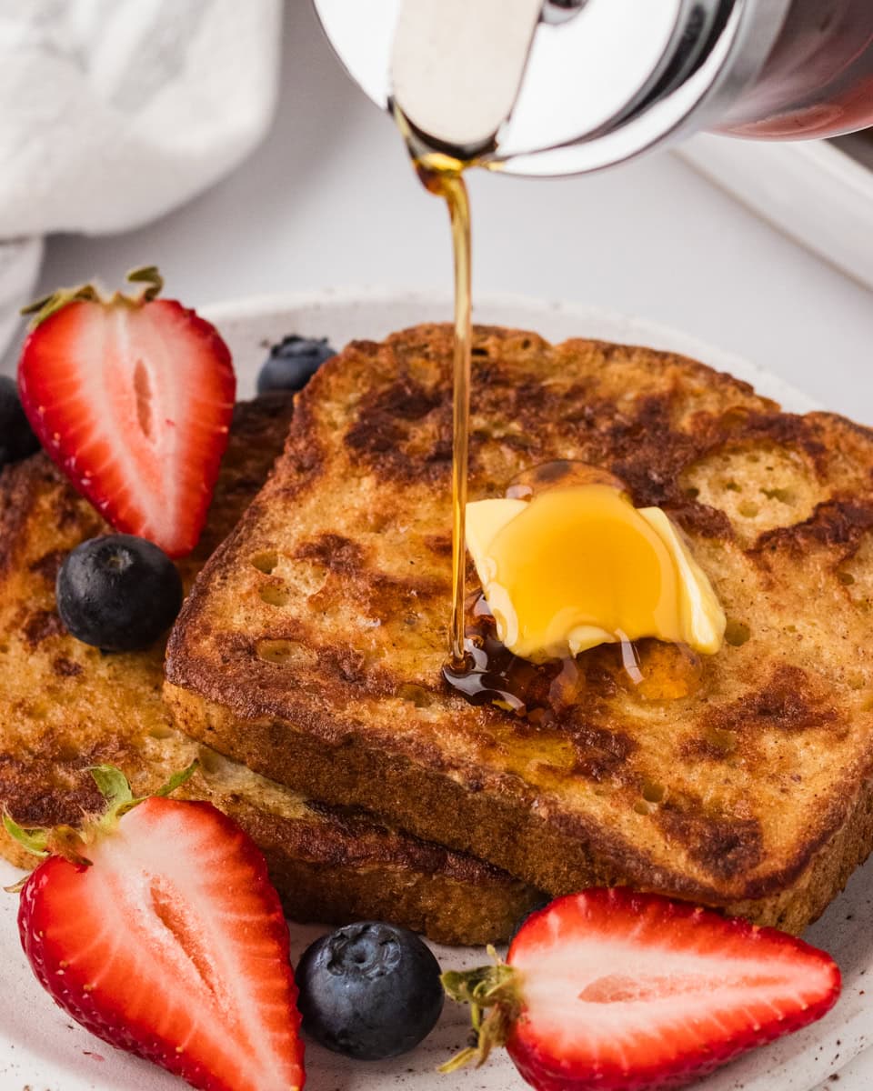 Two slices of golden-brown gluten-free French toast topped with a pat of butter and fresh berries, with maple syrup being poured over the top.