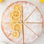 Whole sliced gluten-free olive oil cake topped with powdered sugar and lemon slices, displayed on a marble surface.