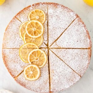 Whole sliced gluten-free olive oil cake topped with powdered sugar and lemon slices, displayed on a marble surface.
