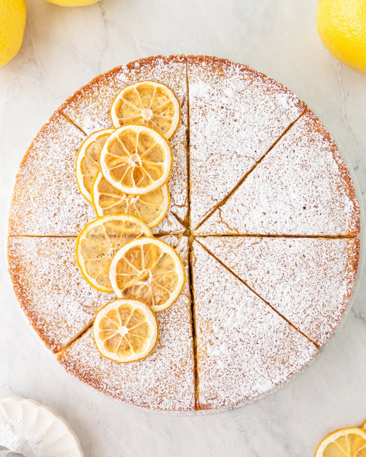 Whole sliced gluten-free olive oil cake topped with powdered sugar and lemon slices, displayed on a marble surface.