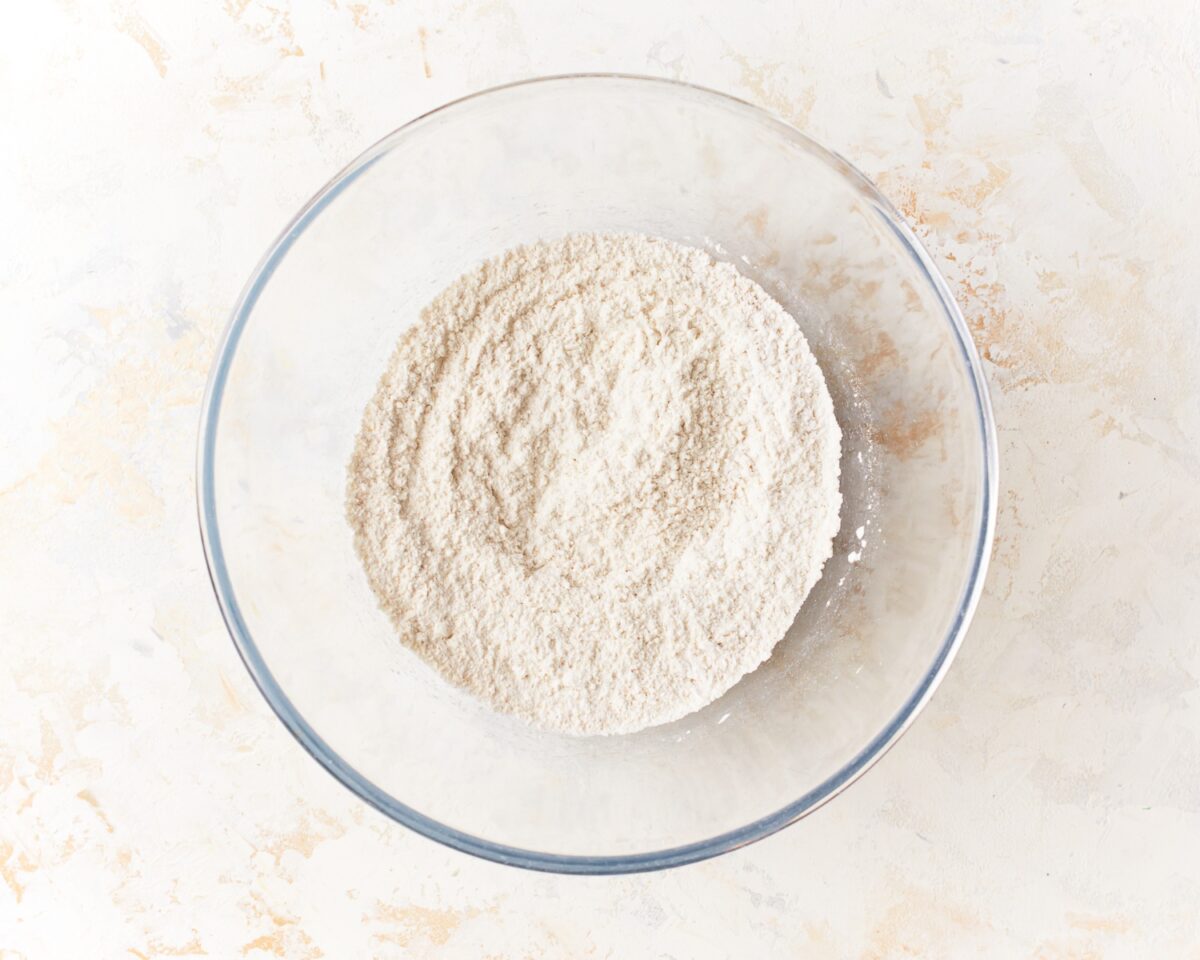 Glass mixing bowl filled with whisked dry ingredients for gluten-free pita bread on a light textured background.