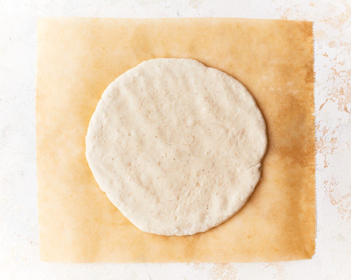 Flattened round of gluten-free pita dough shaped and ready to bake on a sheet of parchment paper.