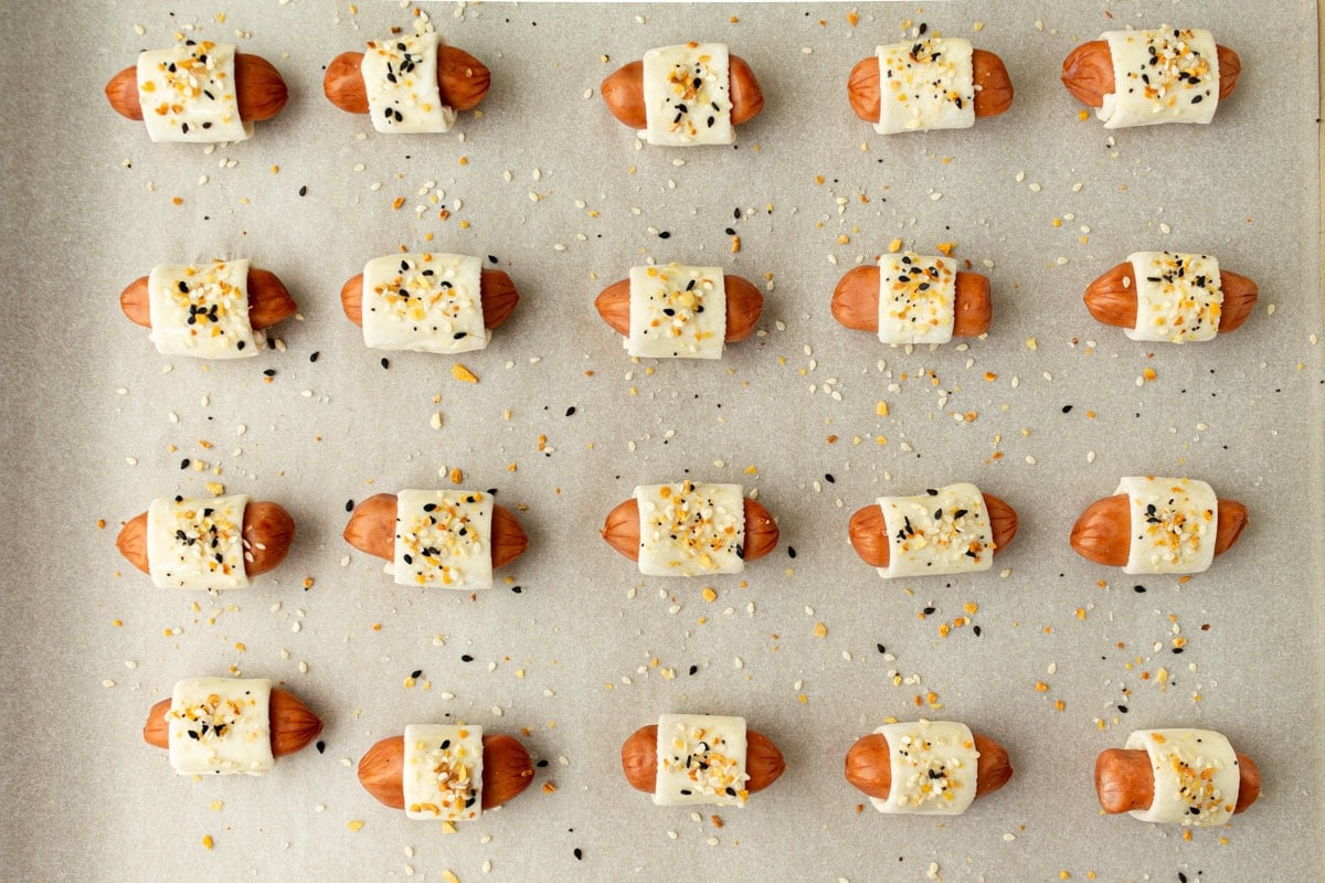 Unbaked pigs in a blanket arranged on a parchment-lined baking sheet, topped with everything bagel seasoning.