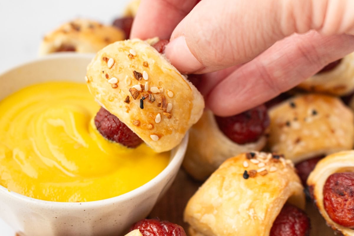 Hand dipping a gluten-free pig in a blanket into a bowl of mustard, surrounded by more on a serving platter.