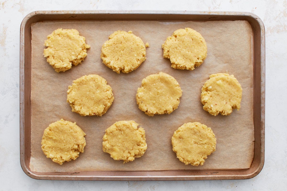 Biscuit dough mounds flattened into round shapes on a baking sheet, ready to bake.