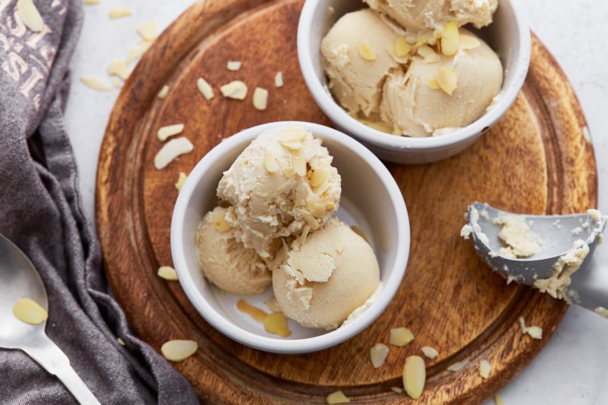 Two ramekins filled with scoops of almond milk ice cream on a wooden board, garnished with sliced almonds and a scoop resting on the side.