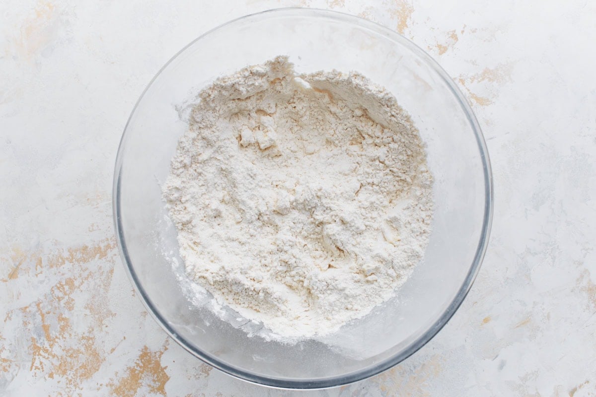 Cold butter cubes cut into the dry mixture, forming a coarse, crumbly dough base in a glass bowl.