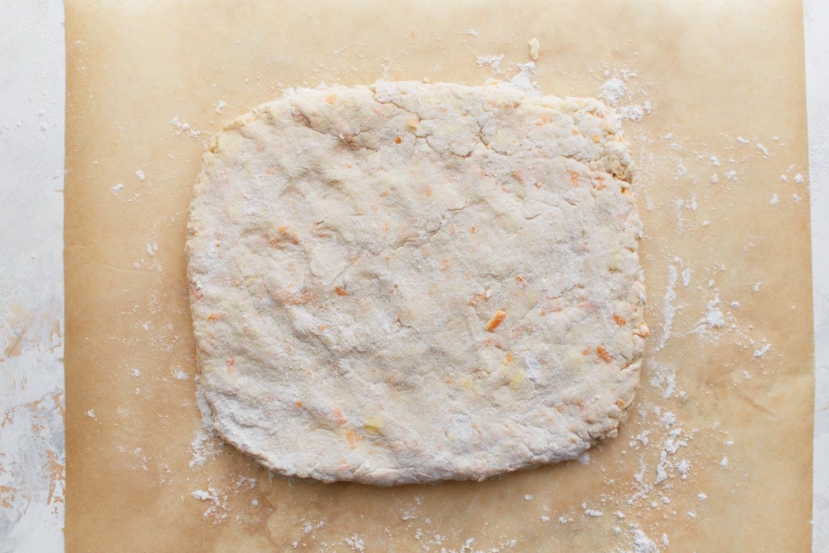 Biscuit dough flattened gently into a rough 1-inch-thick rectangle with visible flour dusting.