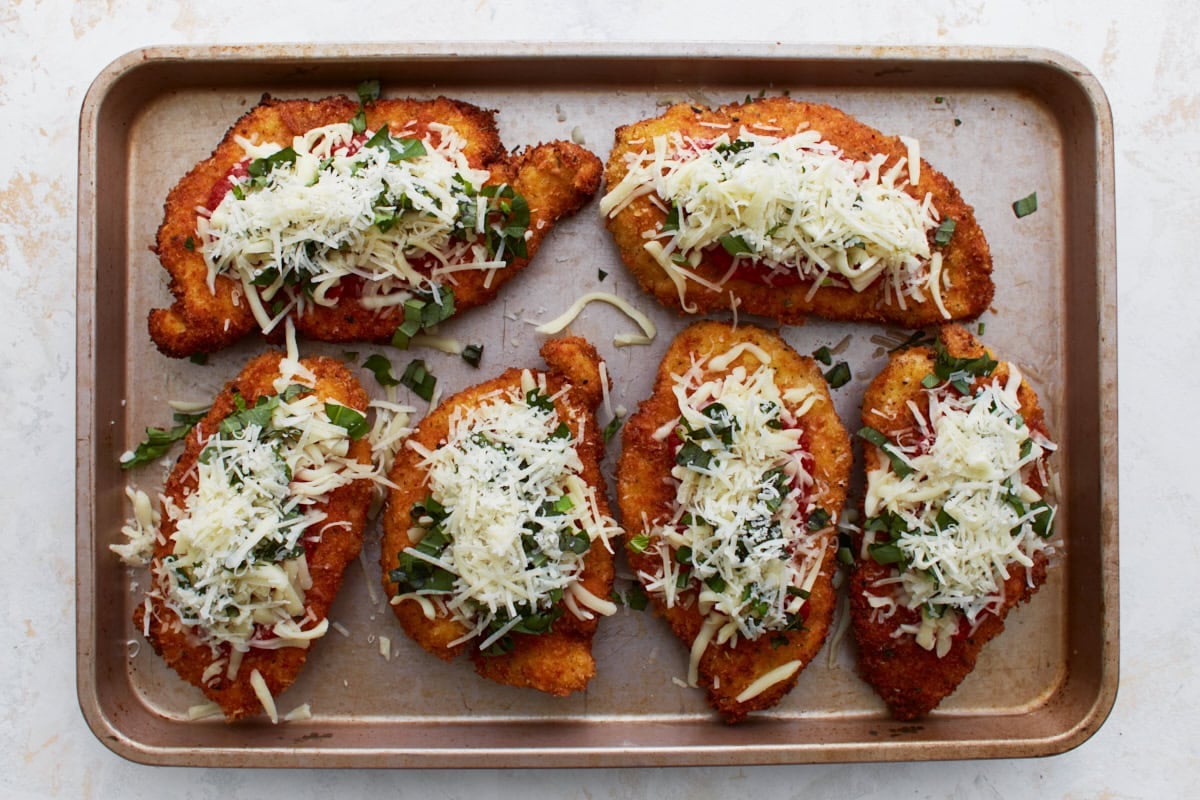 Breaded chicken cutlets topped with shredded mozzarella, chopped basil, and marinara sauce arranged on a baking sheet before baking.