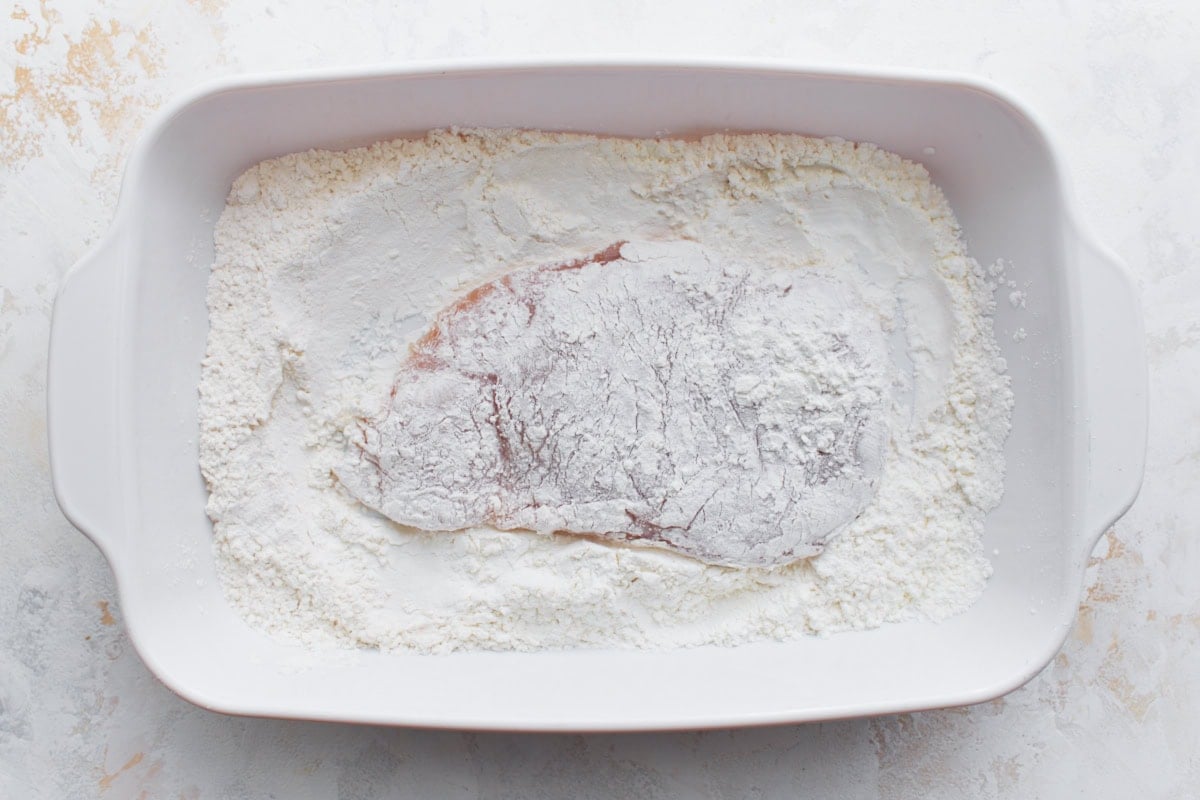 Raw chicken breast dredged in gluten-free flour mixture inside a white baking dish.