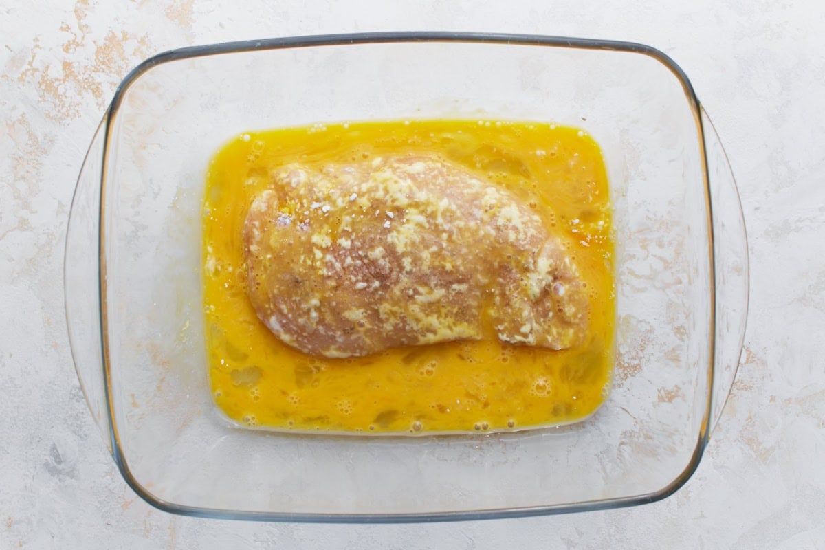 Flour-coated chicken breast dipped in whisked egg mixture inside a glass baking dish.