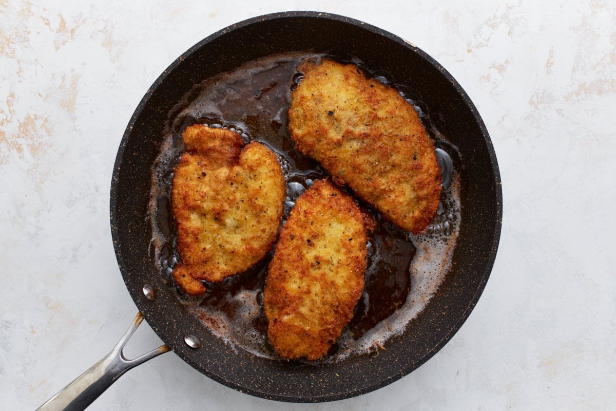 Three breaded chicken breasts pan-frying in hot olive oil inside a skillet.