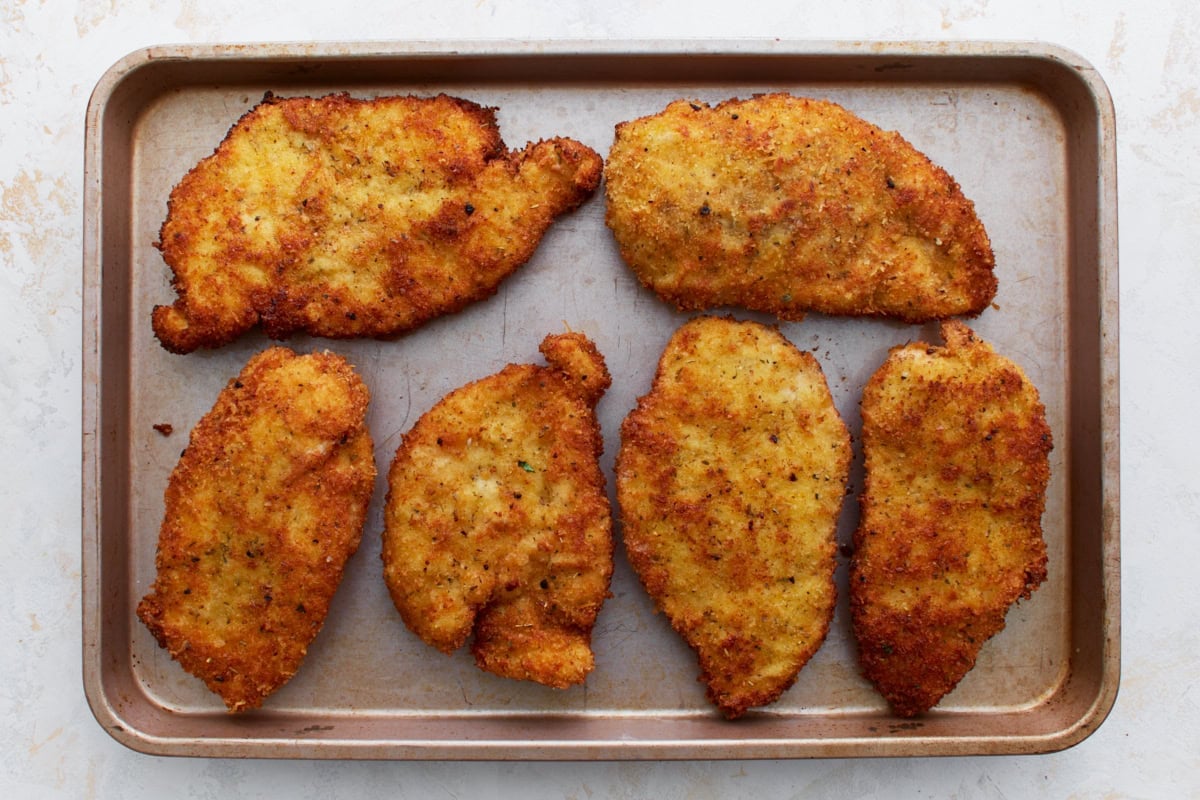 Pan-fried, golden-brown gluten-free chicken breasts arranged on a baking sheet, ready to be topped and baked.