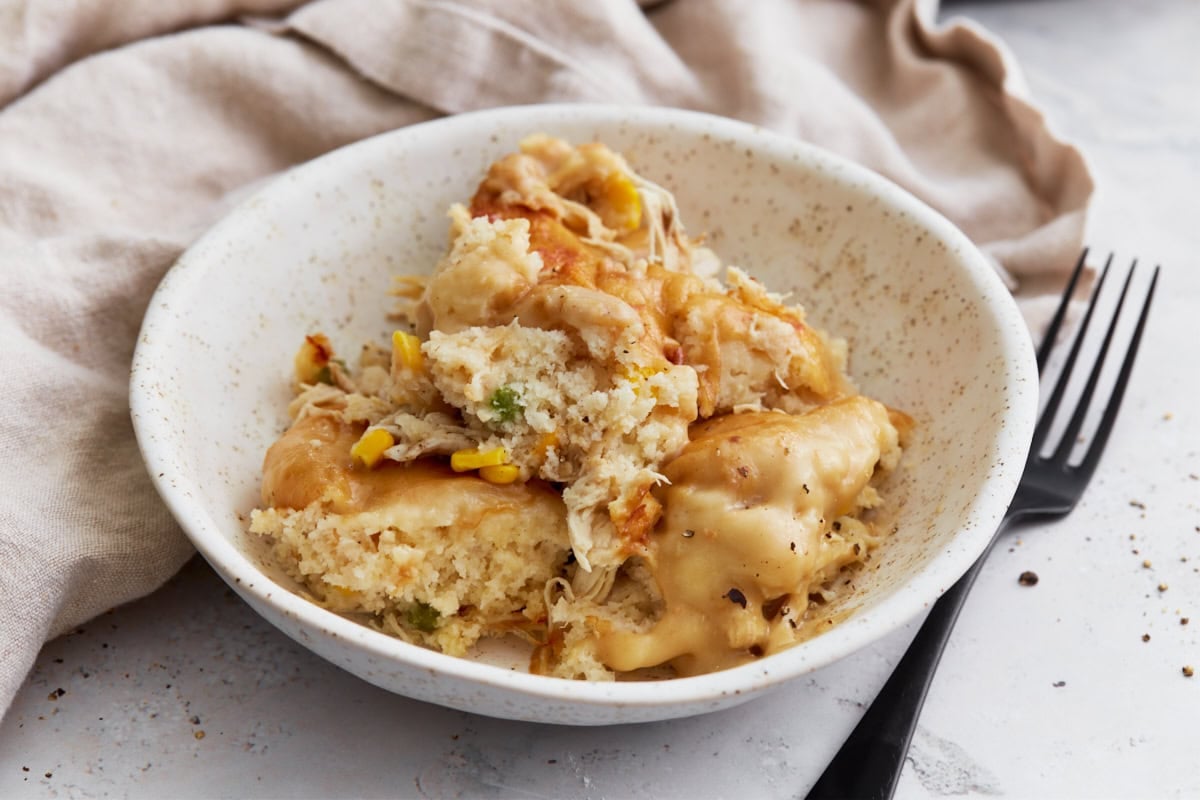 A close-up of a serving of gluten-free chicken and dumplings casserole in a speckled ceramic bowl, showing fluffy dumplings, shredded chicken, corn, and creamy sauce, with a black fork and linen napkin beside it.