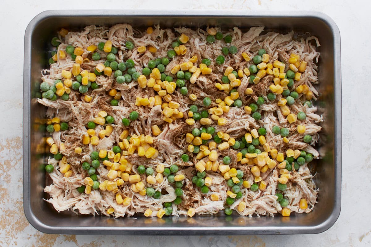 Shredded chicken layered in a baking pan and topped with frozen peas and corn, seasoned with herbs and spices.