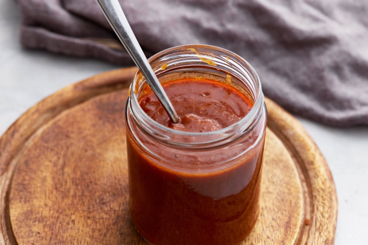 Side view of a glass jar of enchilada sauce on a round wooden board with a spoon inside.