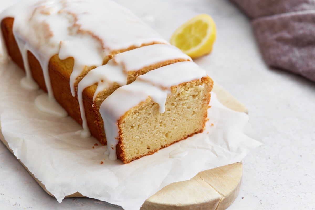 Sliced gluten-free iced lemon loaf on parchment paper, with a lemon half in the background and glaze dripping down the sides.