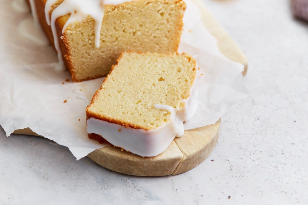 A single slice of gluten-free iced lemon loaf laid flat on parchment, showing the moist crumb and thick glaze along the edges.