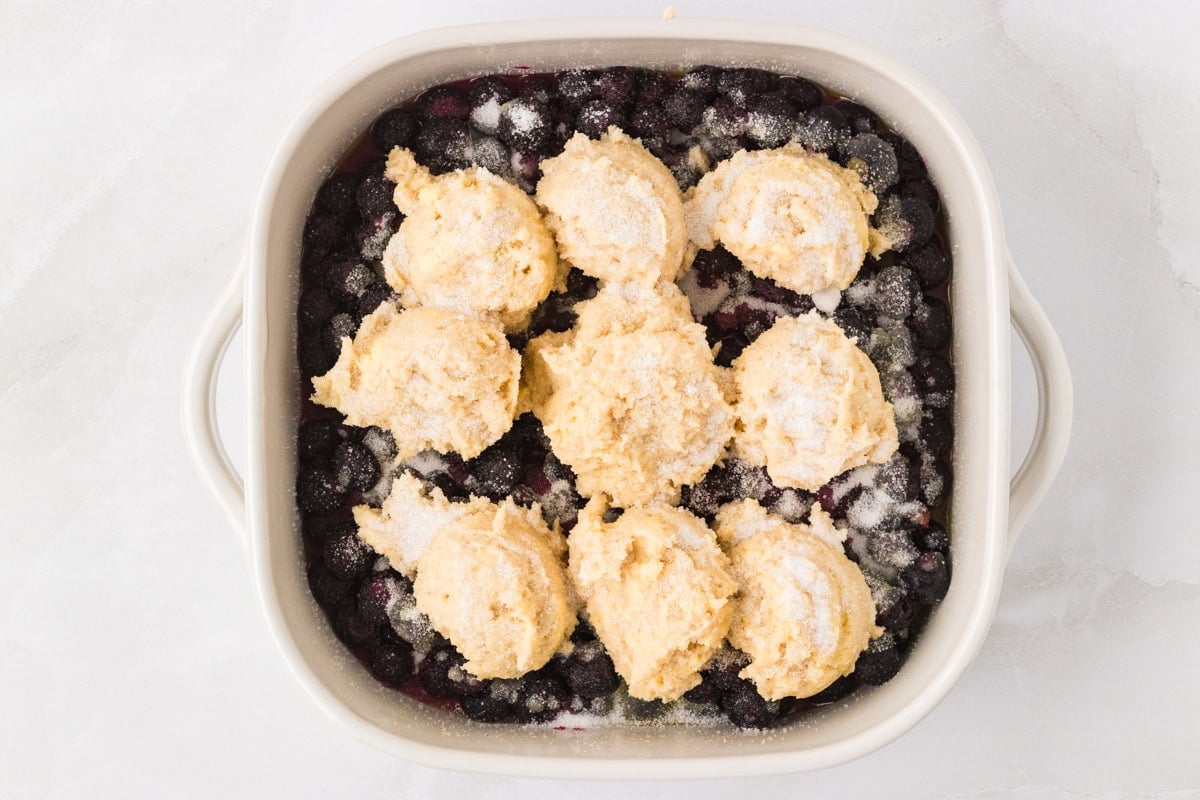 Scoops of biscuit dough placed over the blueberry filling in a baking dish, sprinkled with sugar before baking.