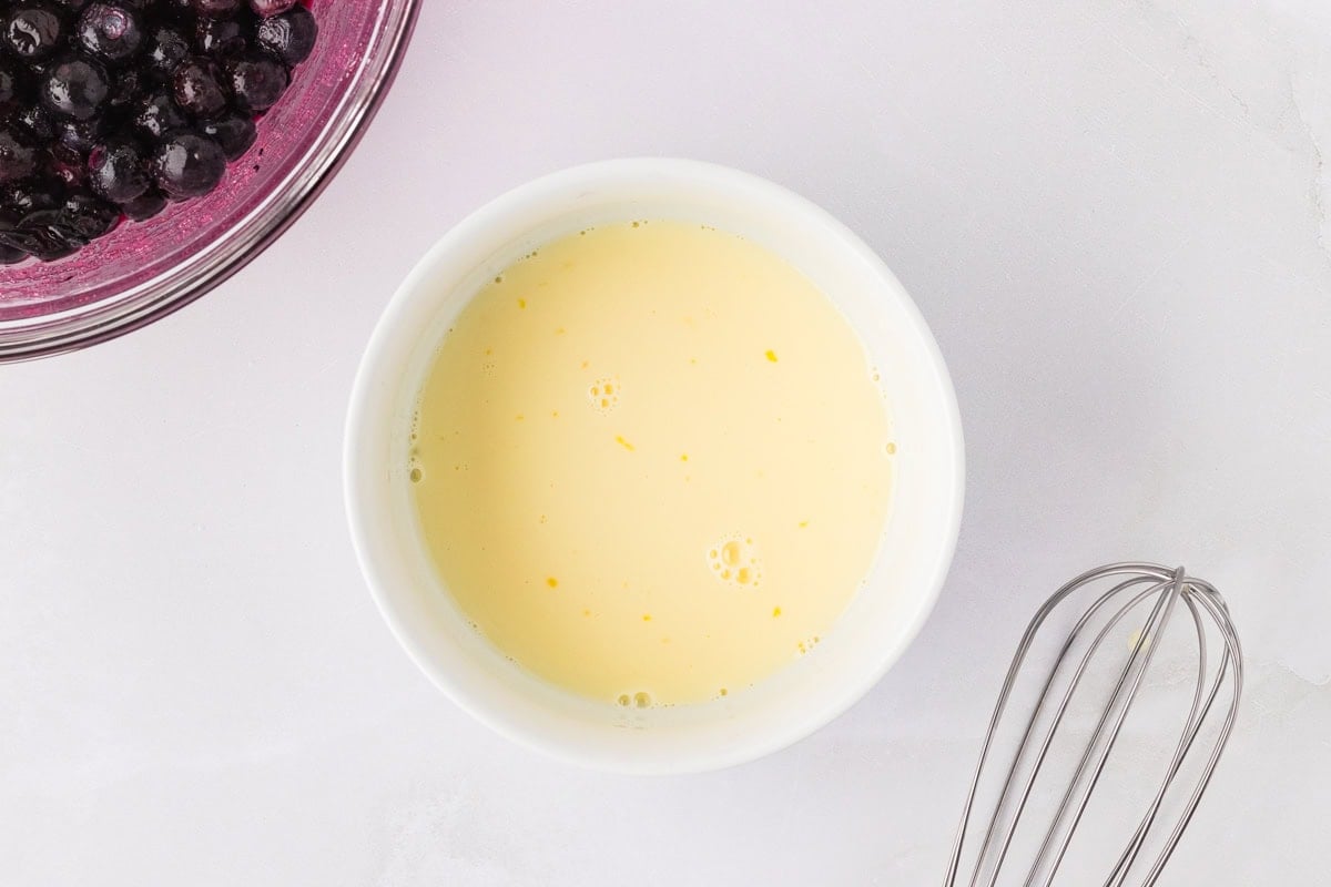 Whisked egg and dairy-free milk in a small white bowl next to a whisk and bowl of blueberries.
