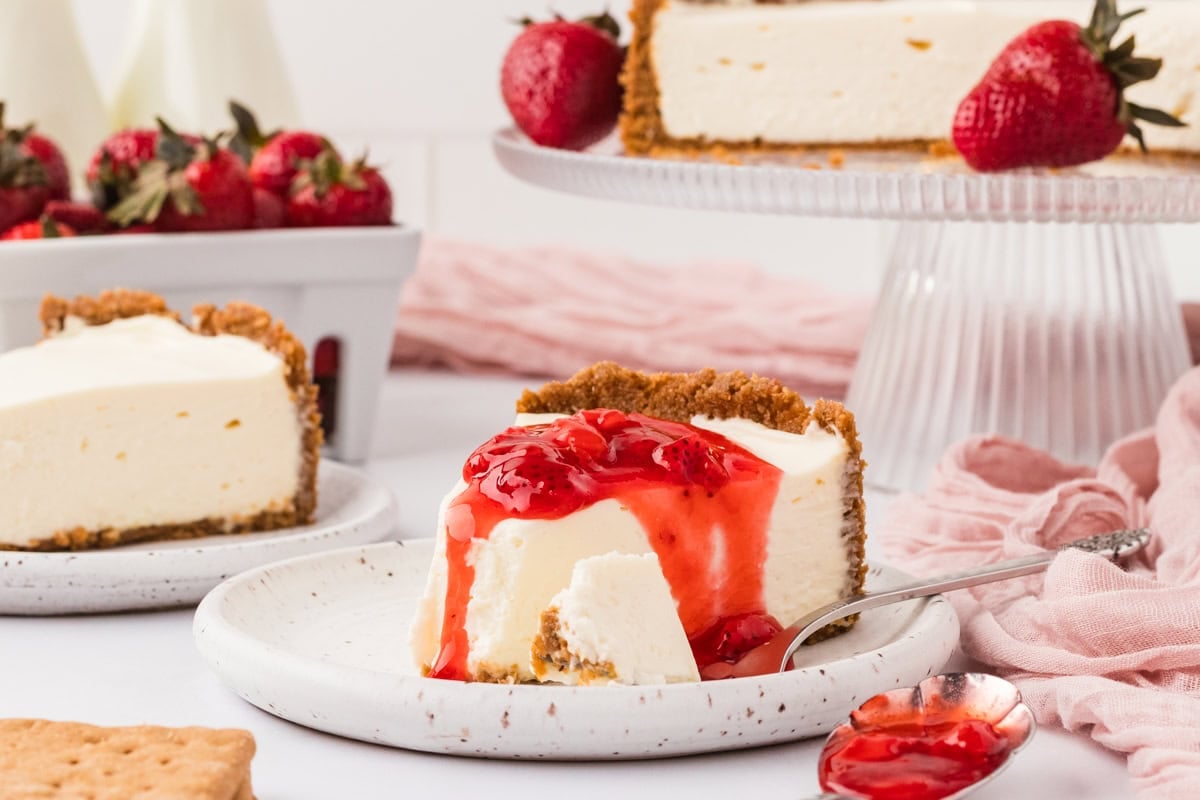 A slice of gluten-free no-bake cheesecake topped with strawberry sauce, with the whole cheesecake and fresh strawberries in the background on a white and pink styled table.
