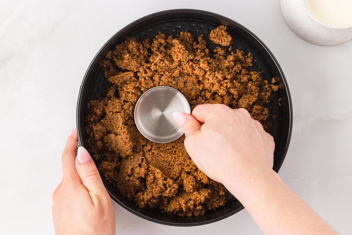 Hands pressing the graham cracker crust into a springform pan using a metal measuring cup.