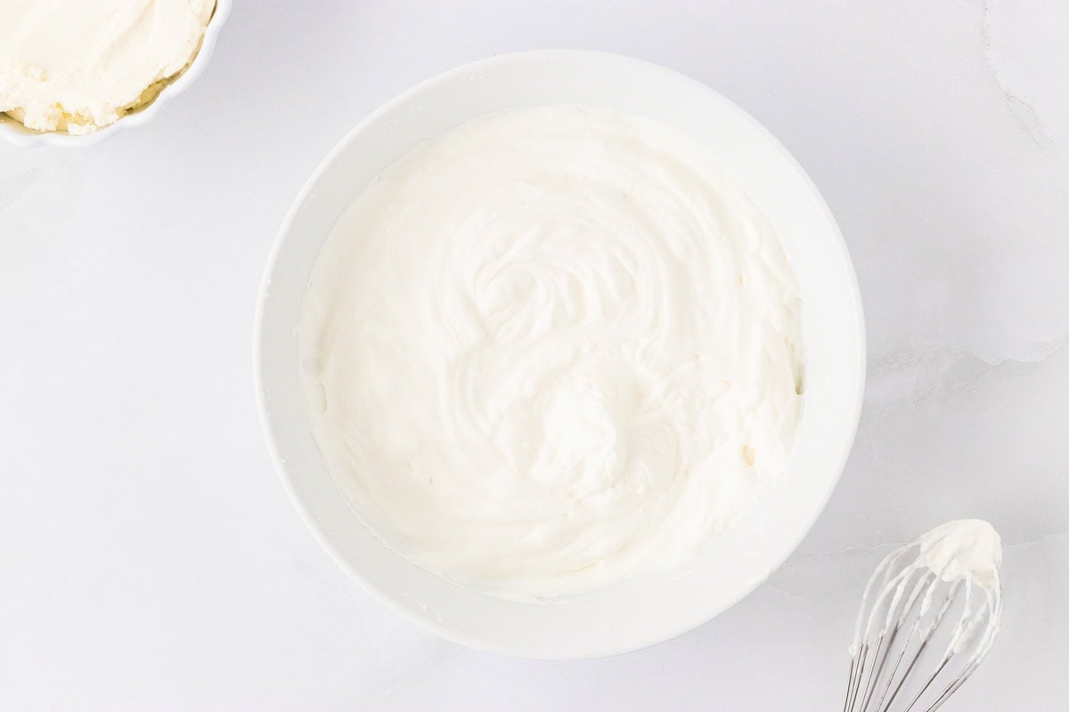 A bowl of freshly whipped cream with soft peaks, ready to be folded into the cheesecake filling.