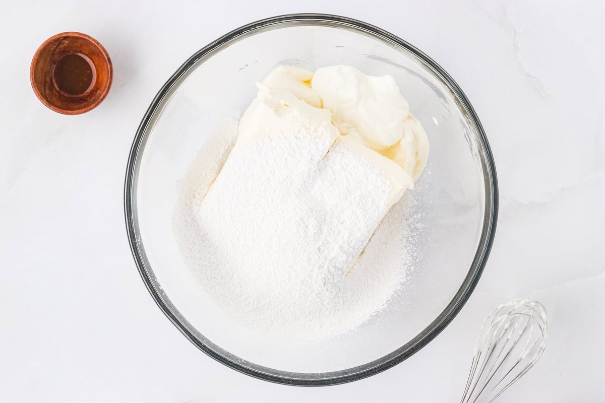 Cream cheese, sour cream, and powdered sugar in a mixing bowl before blending, with a whisk nearby.