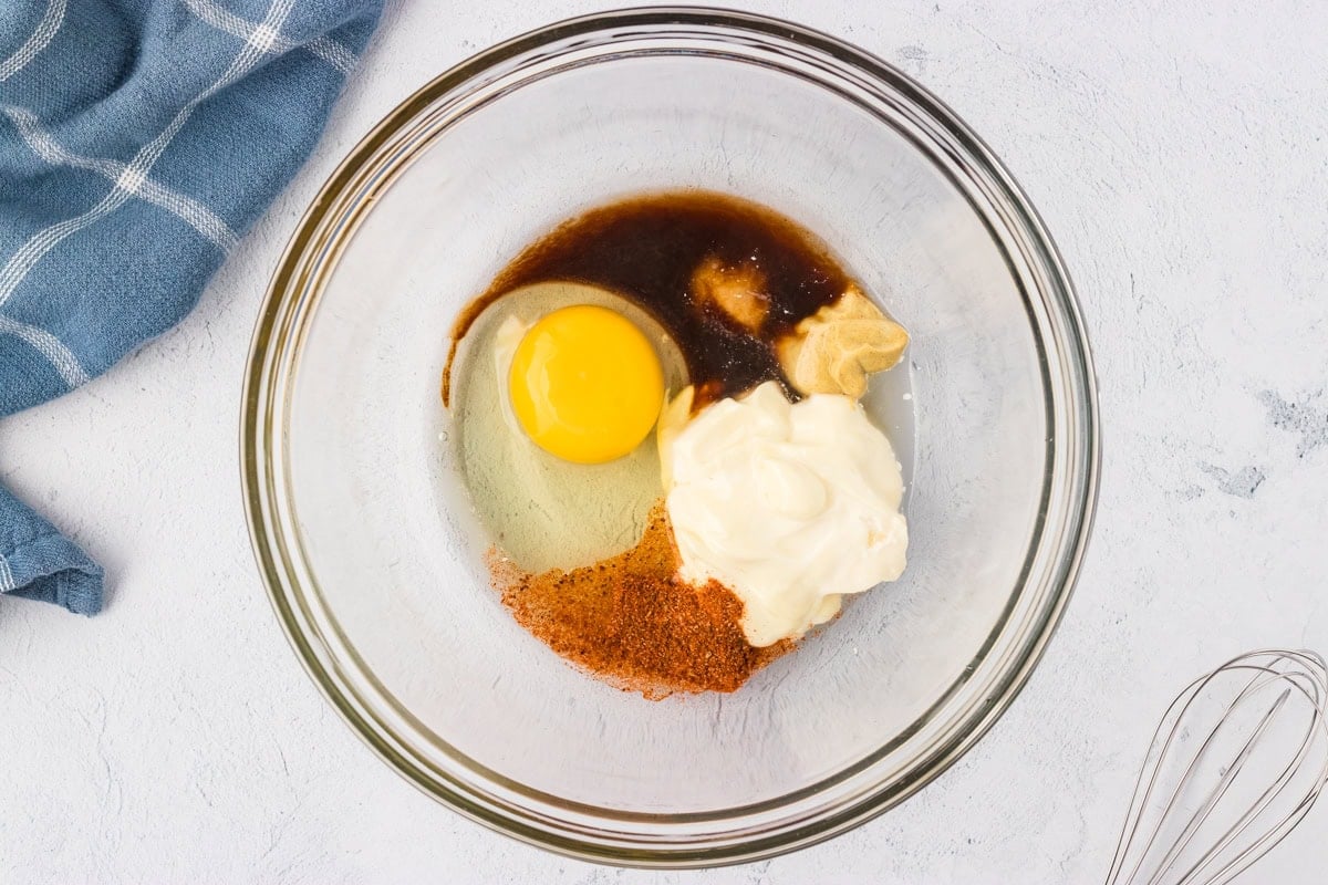A glass mixing bowl with egg, mayonnaise, Dijon mustard, Worcestershire sauce, and seasonings ready to be whisked together.