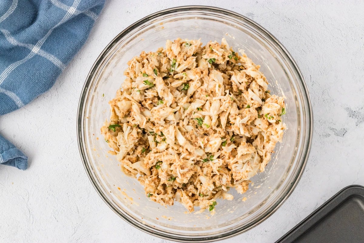 Crab cake mixture fully combined in a glass bowl, showing large chunks of crab and flecks of parsley.