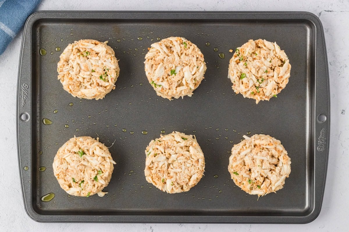 Six unbaked crab cakes formed into mounds on an oiled baking sheet, ready to cook.